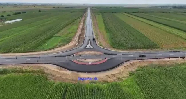 O vedere aeriană prezintă o intersecție rutieră cu un sens giratoriu situată în mijlocul unor câmpuri agricole verzi și maronii.