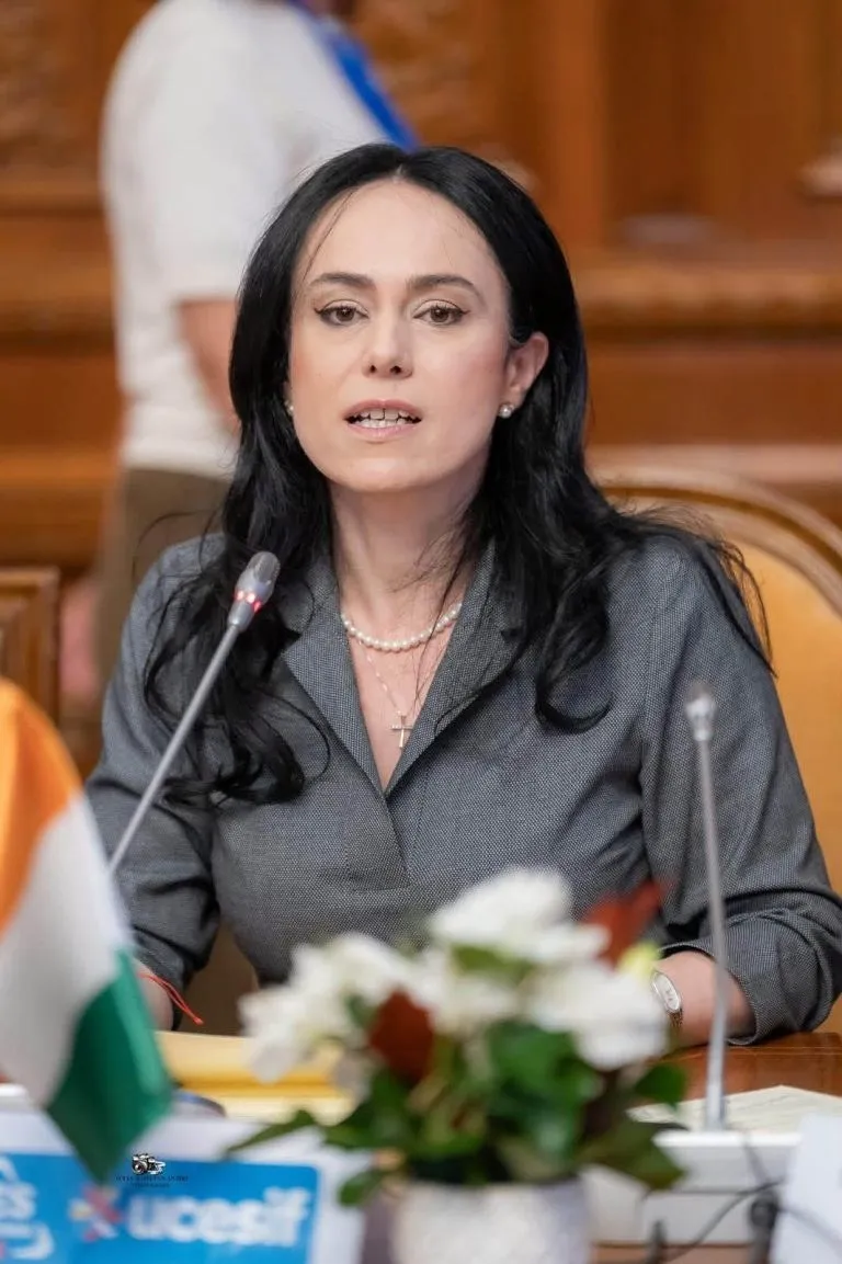 O femeie cu părul negru, purtând o cămașă gri și colier, stă la o masă lângă un microfon și flori.