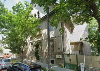 O clădire cu fațadă deschisă, parțial acoperită de copaci verzi, este situată lângă un stâlp de utilități și un gard metalic verde.
