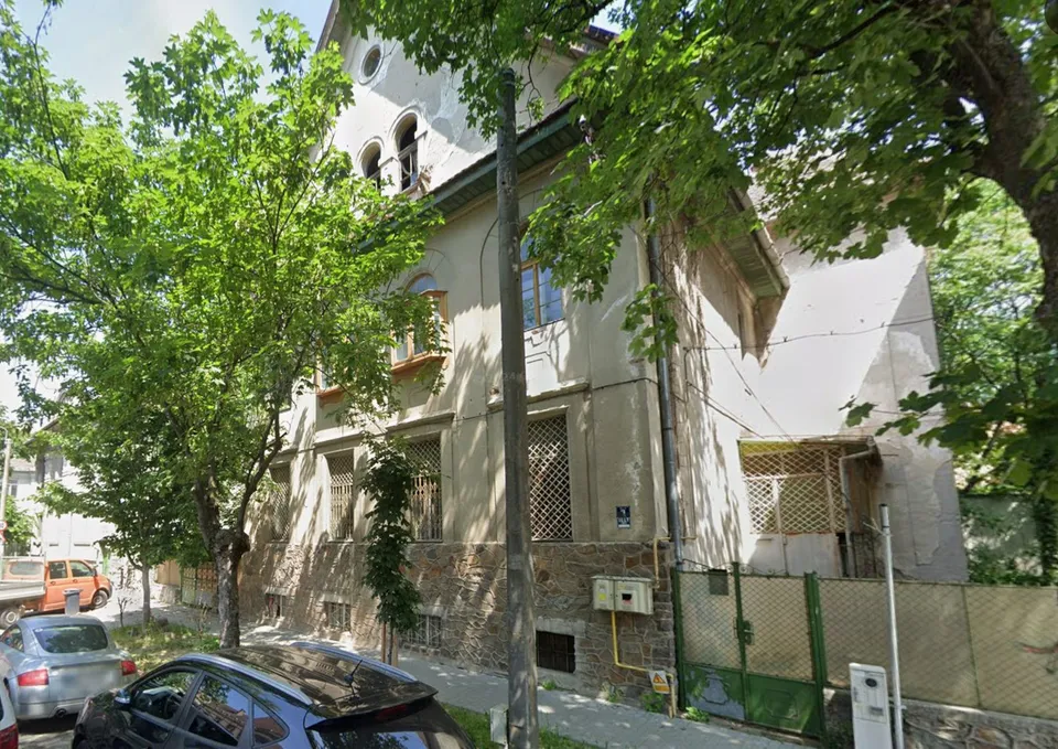 O clădire cu fațadă deschisă, parțial acoperită de copaci verzi, este situată lângă un stâlp de utilități și un gard metalic verde.