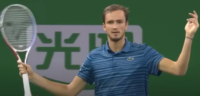 Un bărbat cu tricou albastru în dungi deține o rachetă de tenis și ridică mâna stângă în fața unui fundal verde.