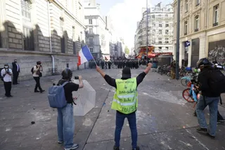 Un bărbat cu un vest vest de culoare galbenă și brațele ridicate stă pe un drum urban lângă alți oameni și biciclete.