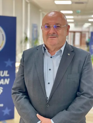 Un bărbat cu ochelari și mustață poartă un sacou gri peste o cămașă deschisă la guler, stând în fața unui fundal albastru cu stele.