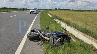 O motocicletă neagră este culcată pe marginea drumului, lângă un șanț și un câmp de cereale, sub un cer cu nori.