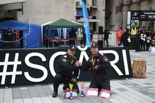 Doi bărbați în uniforme de serviciu negre stau ghemuiți în fața unui panou negru cu textul alb, având trofee și diplome pe podea.