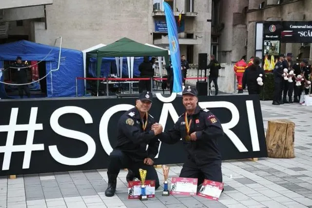 Doi bărbați în uniforme de serviciu negre stau ghemuiți în fața unui panou negru cu textul alb, având trofee și diplome pe podea.