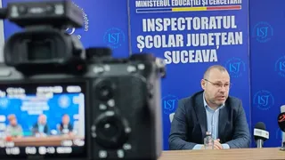 Un bărbat cu ochelari și sacou stă la o masă în fața unui panou albastru cu text alb, fiind filmat de o cameră video în prim-plan.