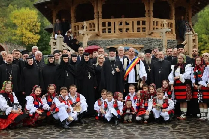 Un grup de oameni, inclusiv clérul în haine negre și copii în costume populare albe cu broderii roșii, posează în fața unei construcții din lemn.