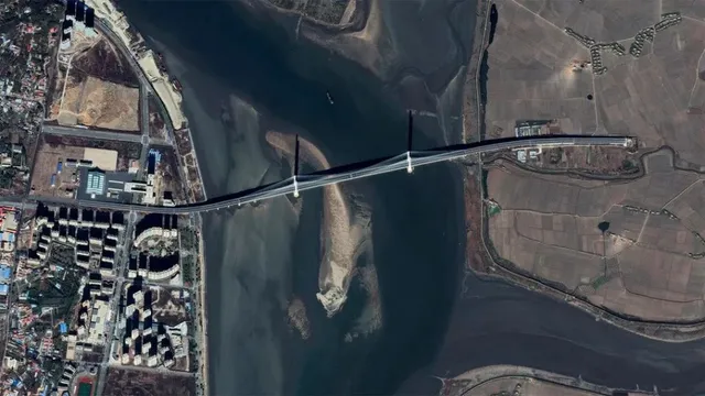 O vedere aeriană prezintă un pod lung care traversează un fluviu larg între zone urbane și terenuri agricole.
