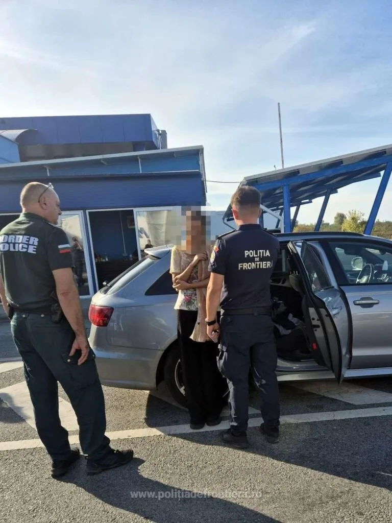 Doi polițiști de frontieră stau lângă o mașină gri, cu o ușă deschisă, și o femeie cu fața blurată, la un punct de trecere a frontierei.