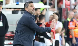 Un bărbat cu părul scurt și barbă gri poartă o jachetă închisă la culoare și stă cu mâinile întinse în față.