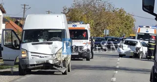 O autoutilitară albă cu avarii mari pe partea dreaptă a capotului este parcată pe marginea drumului, în fundal se observă ambulanțe, mașini de poliție și alte vehicule cu dauni.