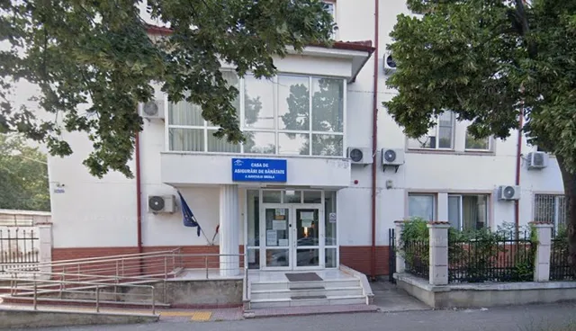 Imaginea prezintă o clădire albă cu ferestre mari, cu o intrare centrală și o rampă de acces. Pe fațadă este un panou albastru cu text alb.