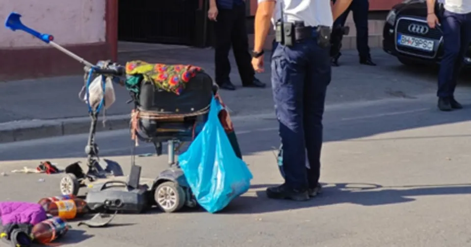 Un scaun rulant electric cu o sacoșă albastră și obiecte împrăștiate pe asfalt stă lângă picioarele unui agent de poliție în uniformă.