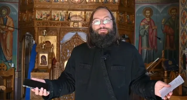 Un bărbat cu barbă și ochelari, îmbrăcat în haine negre, stă în fața unor picturi religioase și ține un obiect negru și o hârtie.