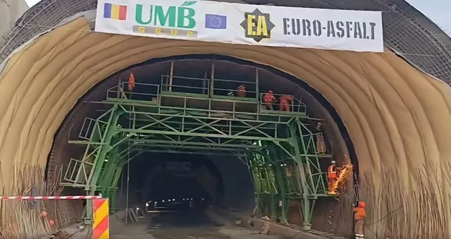 O structură metalică verde de susținere este amplasată în interiorul unei deschideri de tunel, înconjurată de pereți cu finisaj galben și de muncitori în haine portocalii.
