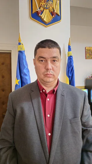 Un bărbat cu păr scurt, purtând sacou gri și cămașă roșie, stă în fața a două steaguri albastre.