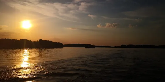 Soarele apune peste un fluviu lat, reflectându-se în apele mișcate sub un cer cu nori subțiri.