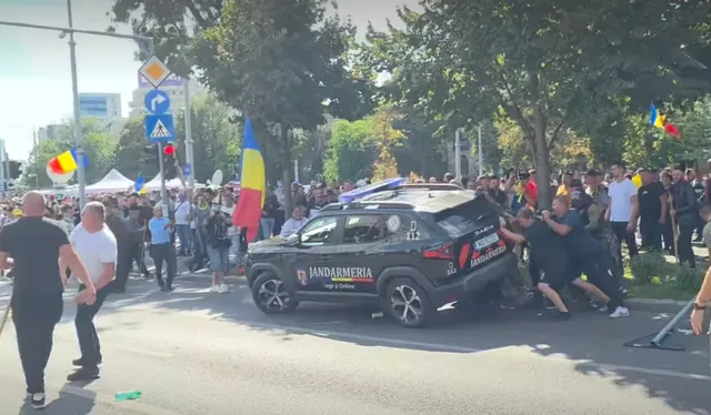 Un grup de persoane în haine închise se prinde de un vehicul negru marcat cu Jandarmeria pe un drum urban cu copaci și steaguri în fundal.