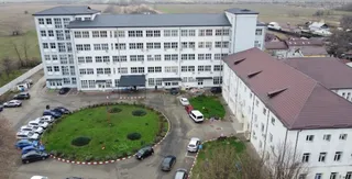 O clădire albă cu mai multe etaje și ferestre dispuse în rânduri este înconjurată de un parcare cu diverse mașini parcate.
