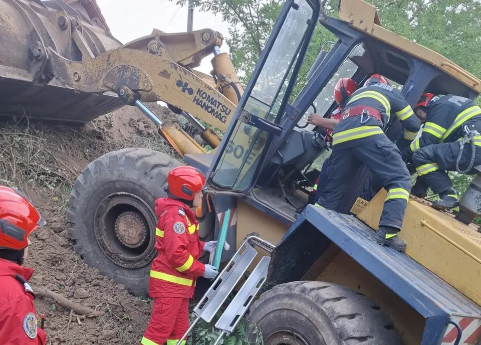 Patru persoane în uniforme de salvator cu accente galbenii și roșii lucrează lângă un utilaj de construcții de culoare galbenă pe un teren accidentat.