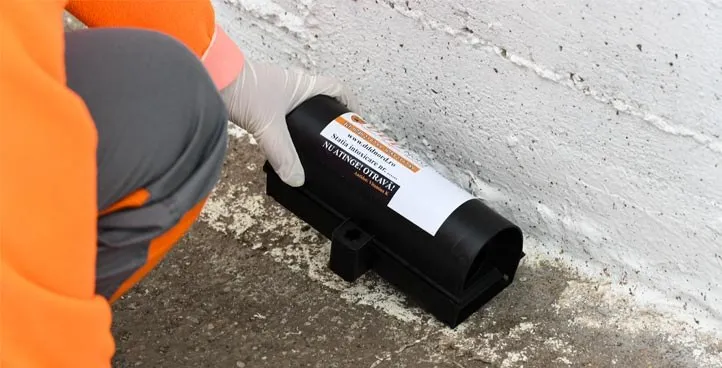O persoană îmbrăcată în haine portocalii și mănuși albe ține un dispozitiv de plastic negru cu un etichetă albă pe un sol de beton lângă un perete alb.