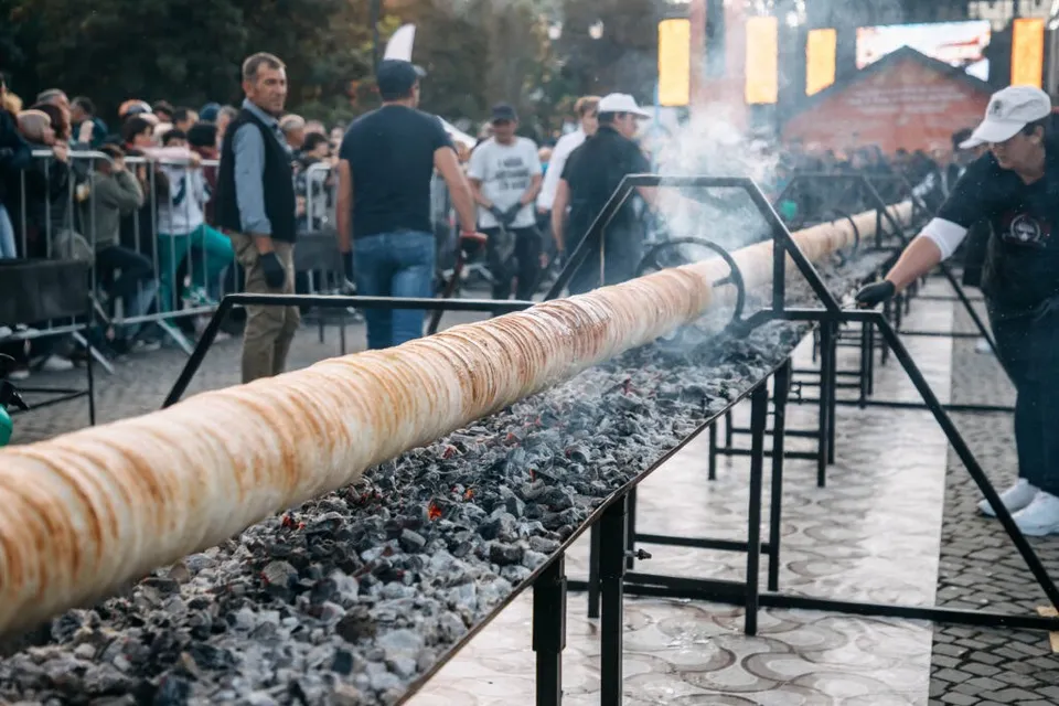 Un colac lung este întins pe un suport metalic deasupra cărbunilor aprinși, în timp ce mai mulți bărbați stau în apropiere.