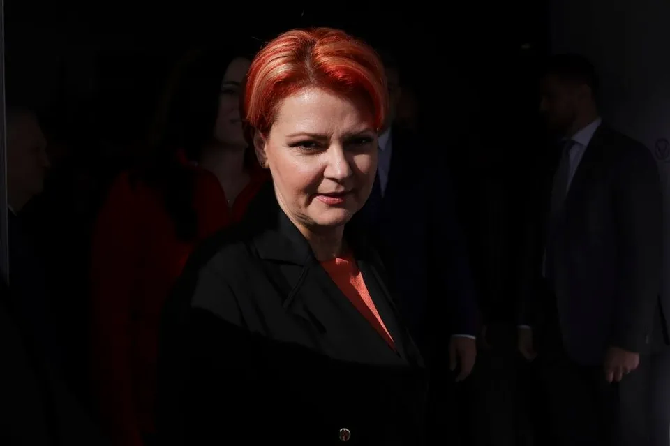 O femeie cu păr roșcat, jachetă neagră și bluză portocalie stă în prim-plan, cu persoane neclare în fundal.