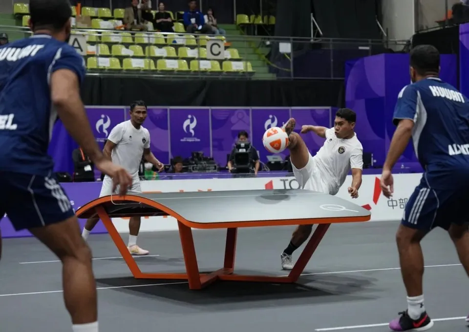 Trei jucători de teqball sunt pe un teren de joc lângă o masă curbată, iar unul dintre ei lovește o minge cu piciorul.