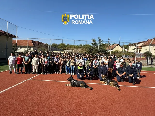 Un grup mare de oameni stă pe un teren sportiv roșu lângă doi câini și doi polițiști în uniformă întunecată.