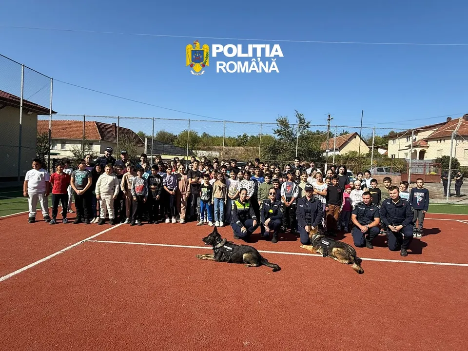Un grup mare de oameni stă pe un teren sportiv roșu lângă doi câini și doi polițiști în uniformă întunecată.