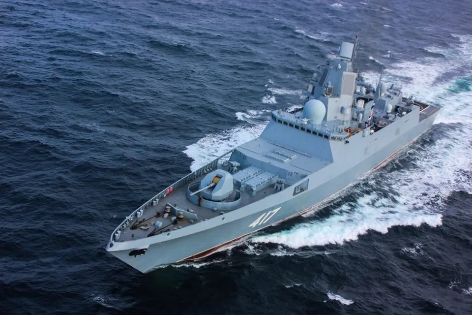 O navă militară gri navigă pe un ocean cu valuri albe, lăsând în urmă o dâră de spumă în urma sa.