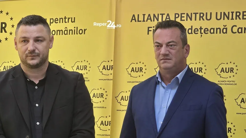 A doi bărbați stau în fața unui fundal galben pe care sunt imprimate diverse logouri negre și text.