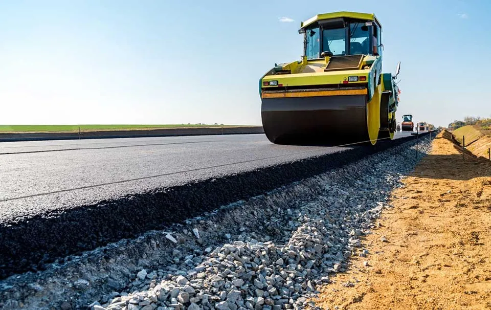 Un aparat de compactare galben rulează pe un strat de asfalt proaspăt întins pe un drum în curs de construcție.