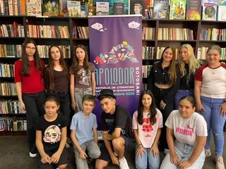 Un grup de adolescenți pozează în fața rafturilor cu cărți și a unui afiș violet, unii stând în picioare, alții ghemuiți.