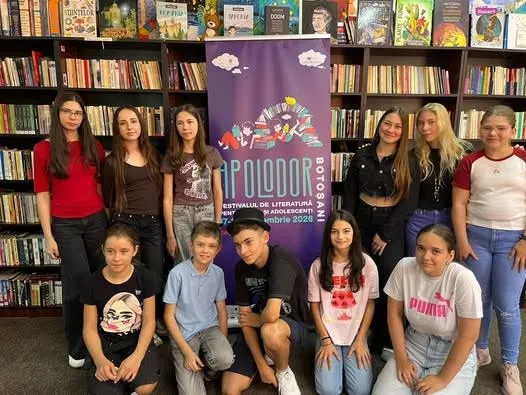 Un grup de adolescenți pozează în fața rafturilor cu cărți și a unui afiș violet, unii stând în picioare, alții ghemuiți.