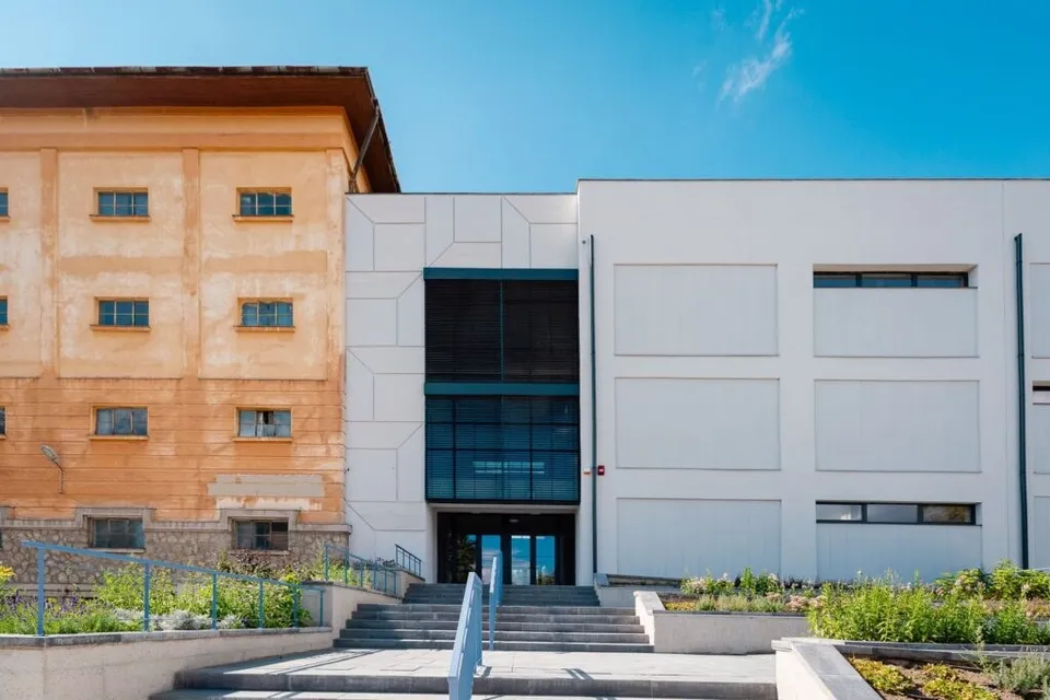 O clădire modernă cu fațadă albă și o secțiune de sticlă neagră este vizibilă lângă o construcție de culoare portocalie sub un cer senin.
