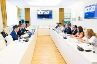 Oamenii stau la o masă lungă, albă, într-o cameră de ședințe cu monitoare afișând imagini pe perete.