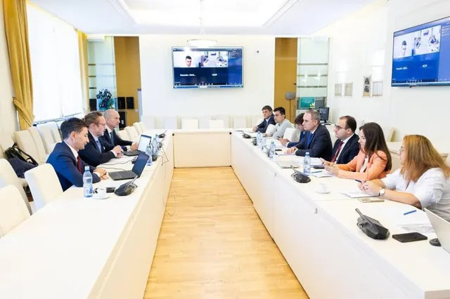 Oamenii stau la o masă lungă, albă, într-o cameră de ședințe cu monitoare afișând imagini pe perete.
