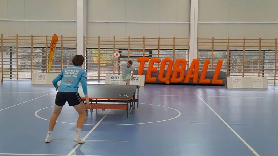 Un bărbat cu tricou albastru și pantaloni scurți negri stă în fața unei mese de teqball pe un teren de sport de culoare albastră.