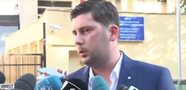 Un bărbat cu părul scurt și sacou albastru vorbește în fața mai multor microfoane de culoare neagră și albastră.