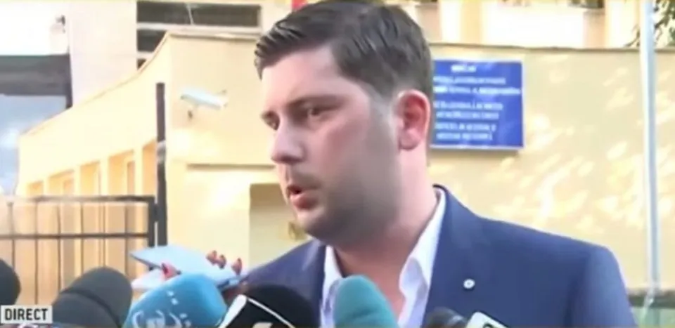 Un bărbat cu părul scurt și sacou albastru vorbește în fața mai multor microfoane de culoare neagră și albastră.