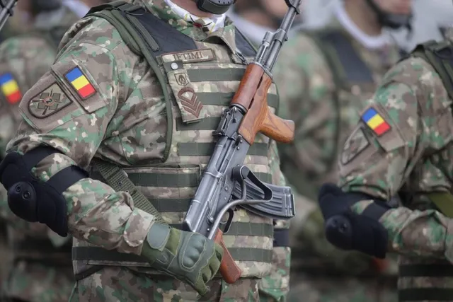 Un soldat în uniformă de camuflaj cu un steag român pe braț ține un pușcă cu coci de lemn.