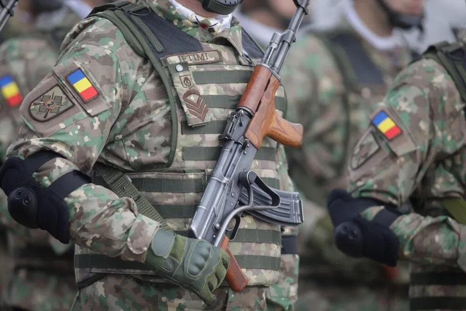 Un soldat în uniformă de camuflaj cu un steag român pe braț ține un pușcă cu coci de lemn.
