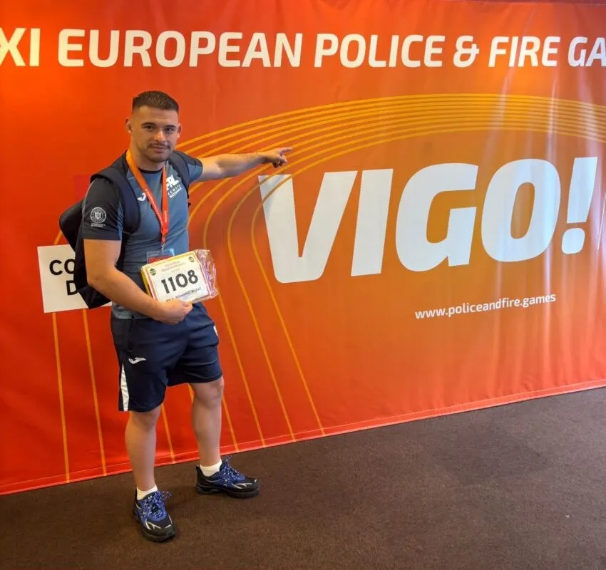 Un bărbat cu tricou albastru și pantaloni scurți poartă o medalie și un număr de concurs în fața unui panou portocaliu cu textul VIGO!.
