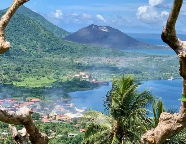 O vedere aeriană a unui oraș de coastă cu clădiri colorate, înconjurat de vegetație luxuriantă, un vulcan și un corp de apă albastru, sub un cer senin.