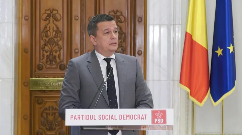 Un bărbat într-un costum gri și cravată neagră stă în spatele unui pupitru alb marcat cu textul PARTIDUL SOCIAL DEMOCRAT și un logo roșu.