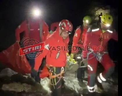 Patru persoane purtând haine roșii și căști de protecție transportă o victimă pe o targă într-un teren accidentat noaptea.