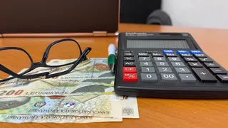 Ochelari cu ramă neagră, un calculator de birou și câteva bancnote de două sute de lei sunt așezate pe o suprafață de lemn.