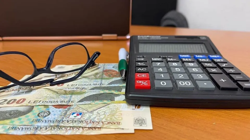 Ochelari cu ramă neagră, un calculator de birou și câteva bancnote de două sute de lei sunt așezate pe o suprafață de lemn.